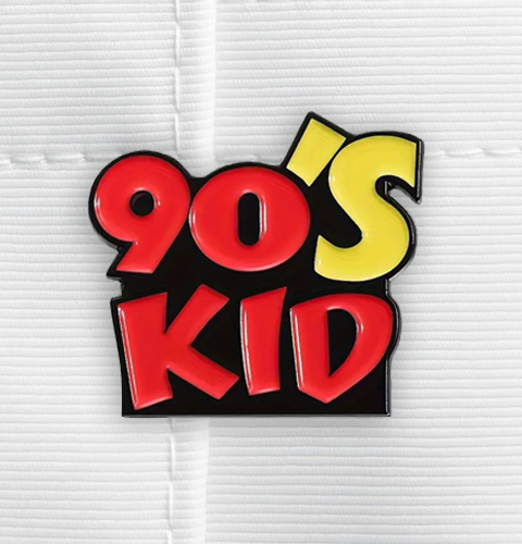Enamel pin with '90's Kid' text in red and yellow on a black border against a white background.