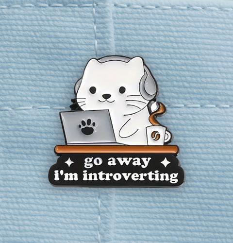 Enamel pin with a cat design and text 'go away I'm introverting' on a blue denim background