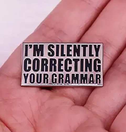 Metal badge with text 'I'm Silently Correcting Your Grammar' held in a hand.
