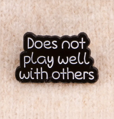 Black enamel pin with white text 'Does not play well with others' on a beige background