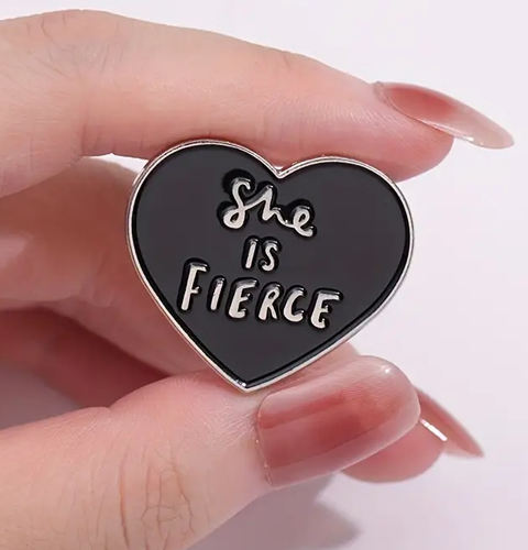 Heart-shaped pin with 'She is Fierce' text held by a hand on a white background