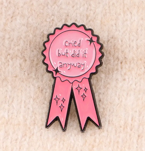 Pink ribbon-shaped pin with text on a beige textured background