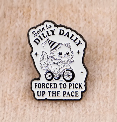 Enamel pin with a cat on a bicycle and text 'Born to Dilly Dally, Forced to Pick Up the Pace' on a beige background
