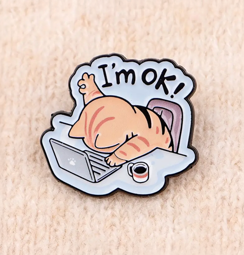 Enamel pin with cartoon cat lying on a laptop and coffee cup, text 'I'm ok!' on a light brown background