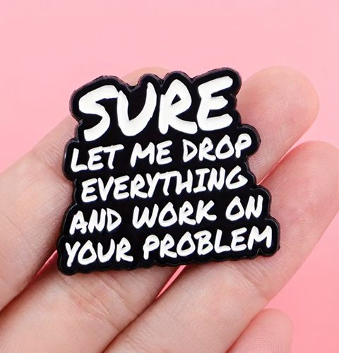 Enamel pin with text 'SURE LET ME DROP EVERYTHING AND WORK ON YOUR PROBLEM' held in a hand against a pink background